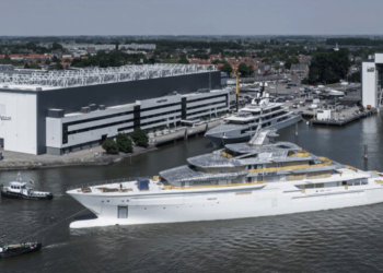 Oceano's Y722 project reaches a significant milestone, MTB Events, Image shows the Y722 superyacht being transported by three smaller work vessels in a port alongside two large buildings labelled 'Oceano', the water s dark and the sky is clear