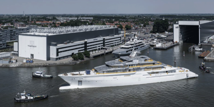Oceano's Y722 project reaches a significant milestone, MTB Events, Image shows the Y722 superyacht being transported by three smaller work vessels in a port alongside two large buildings labelled 'Oceano', the water s dark and the sky is clear