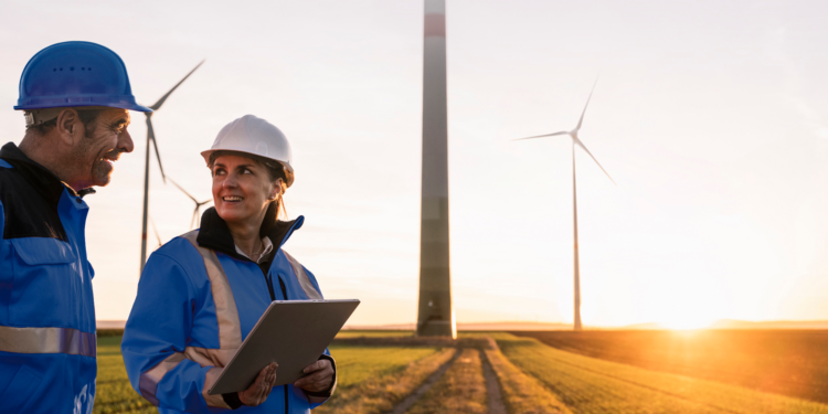 RWE wins tender for 30MW wind farm in Hessian, MTB Events, Image shows a man and woman looking at a clipboard at a wind farm at sunset, both persons are in blue safety jackets and helmets