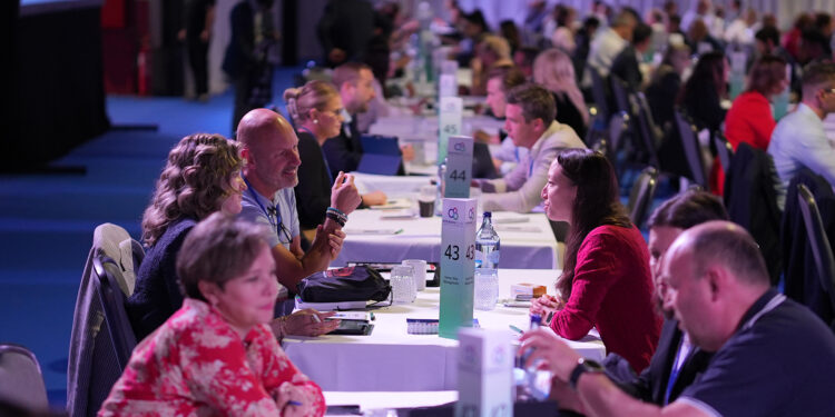 Image shows white tables going vertically along a hall with people sat either side in a room lit with blue and pink lights. Connections, Collaboration and Community was the theme of this year’s MTB Marine Europe , MTB Events.